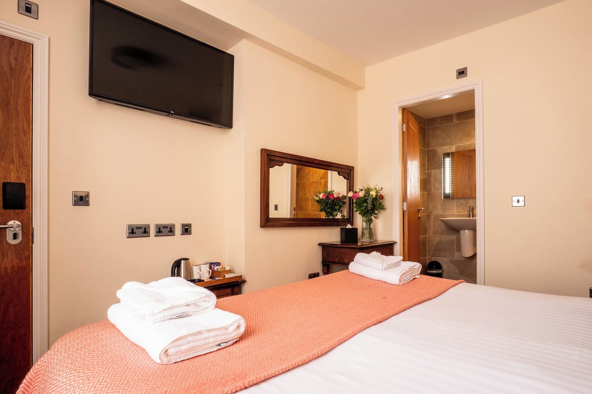 Ein Hotelzimmer mit einem Bett, einem an der Wand befestigten Fernseher, einem Spiegel, einer Vase mit Blumen und einem durch eine offene Tür einsehbaren Badezimmer.