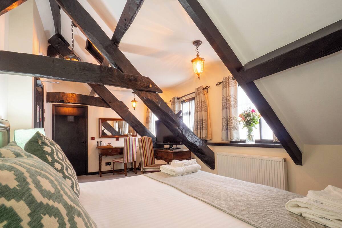 Ein gemütliches Schlafzimmer im Dachgeschoss mit sichtbaren Holzbalken, einem Bett mit einem gemusterten Kissen, einem Schreibtisch mit Stuhl und einem Fenster mit Vorhängen.