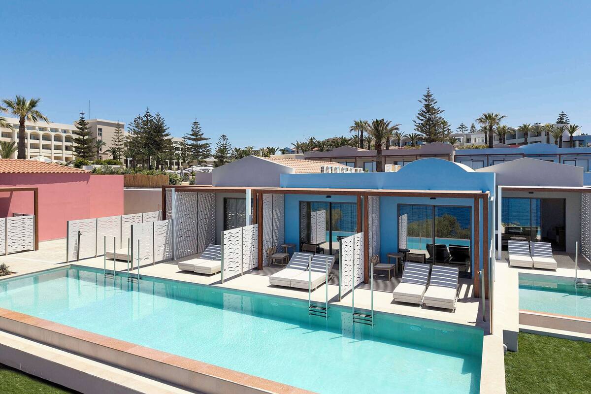 Un espace piscine doté de chaises longues et d’un coin salon abrité, le tout entouré de bâtiments et de palmiers.