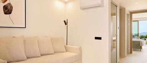 A modern hotel room with a beige sofa, a framed artwork, an air conditioning unit, and a view of the outdoors.
