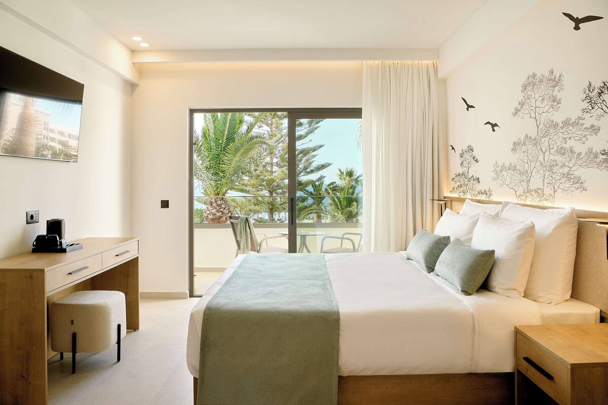 Une chambre d’hôtel moderne dotée d’un grand lit, d’un bureau et d’un balcon offrant une vue sur les palmiers.