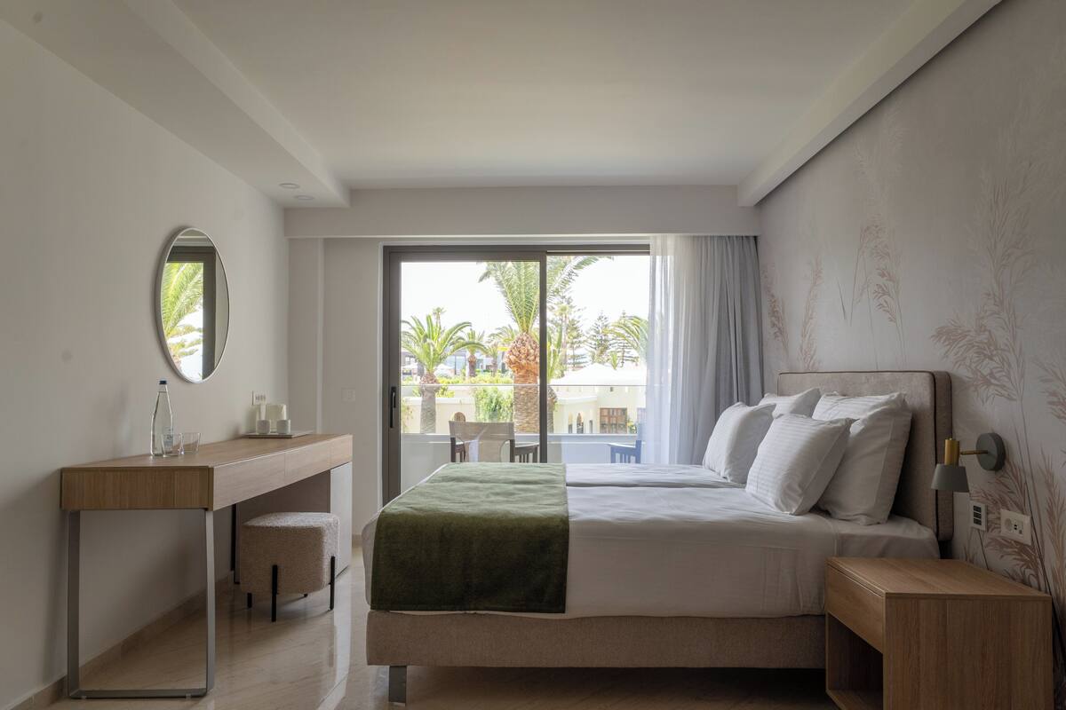 Une chambre d’hôtel avec un lit, un bureau, une chaise, un petit tabouret, un miroir et une vue sur les palmiers et les immeubles, visible à travers une grande fenêtre.
