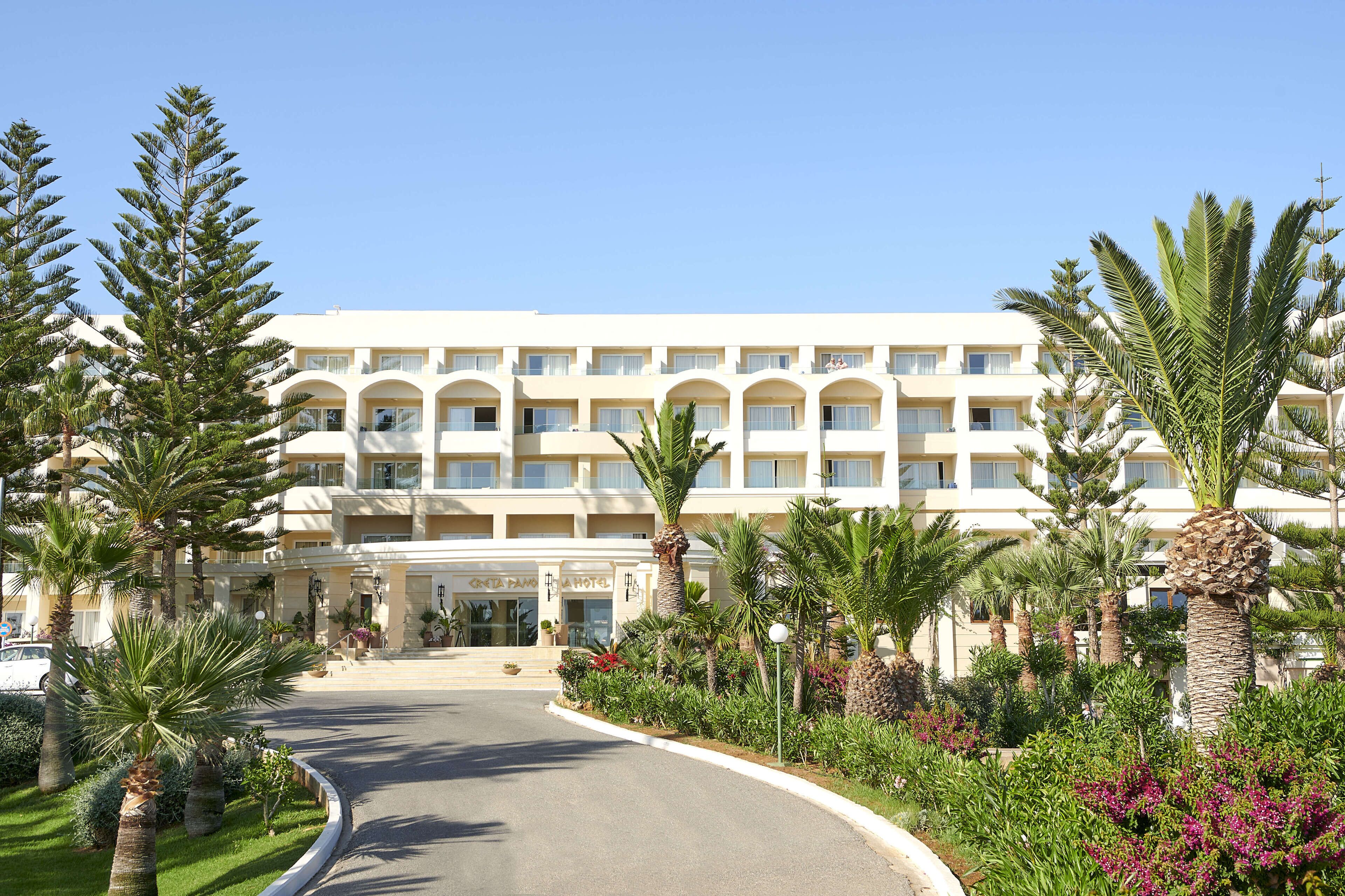 Ein mehrstöckiges Hotel mit einem zentralen Eingang, umgeben von Palmen und verschiedenen Pflanzen.