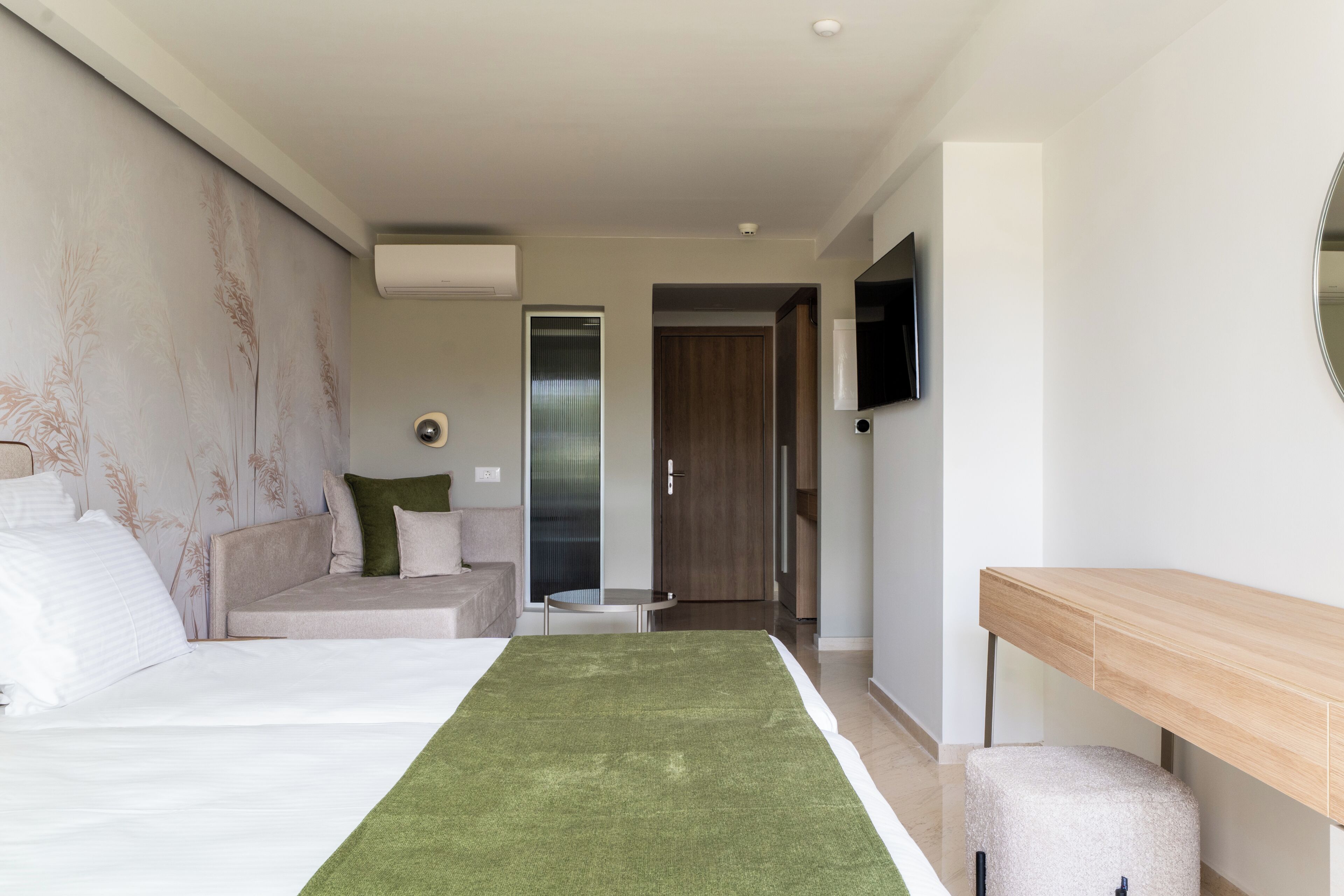 A hotel room with a bed, a green area rug, a wooden bench, a TV mounted on the wall, and a door leading to another room.