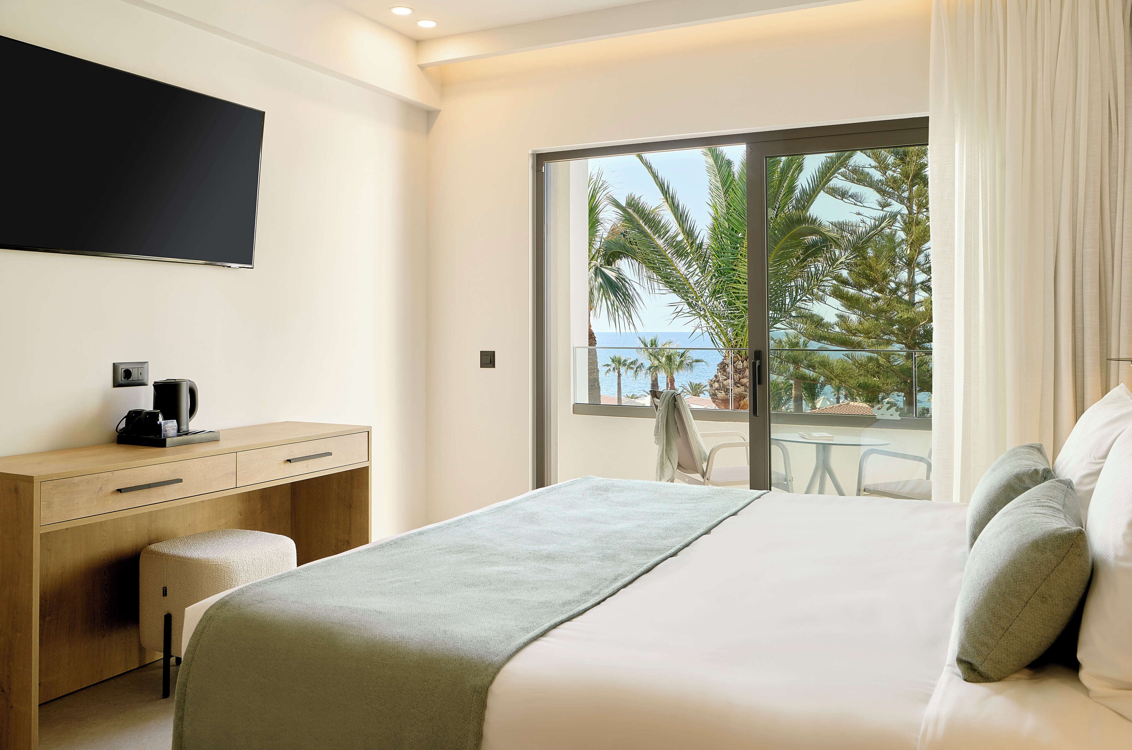 A modern hotel room with a large bed, a desk with a coffee maker, a flat-screen TV, and a balcony with a view of palm trees and the sea.