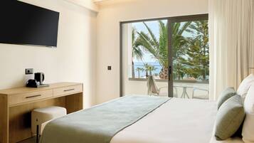 A modern hotel room with a large bed, a desk with a coffee maker, a flat-screen TV, and a balcony with a view of palm trees and the sea.