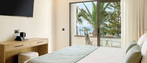 Ein modernes Hotelzimmer mit einem großen Bett, einem Schreibtisch mit Kaffeemaschine, einem Flachbildfernseher und einem Balkon mit Blick auf Palmen und das Meer.