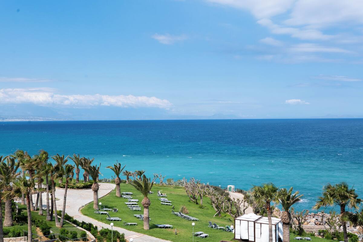 Un hôtel situé sur la côte, offrant une vue sur la mer, des palmiers et des chaises longues.