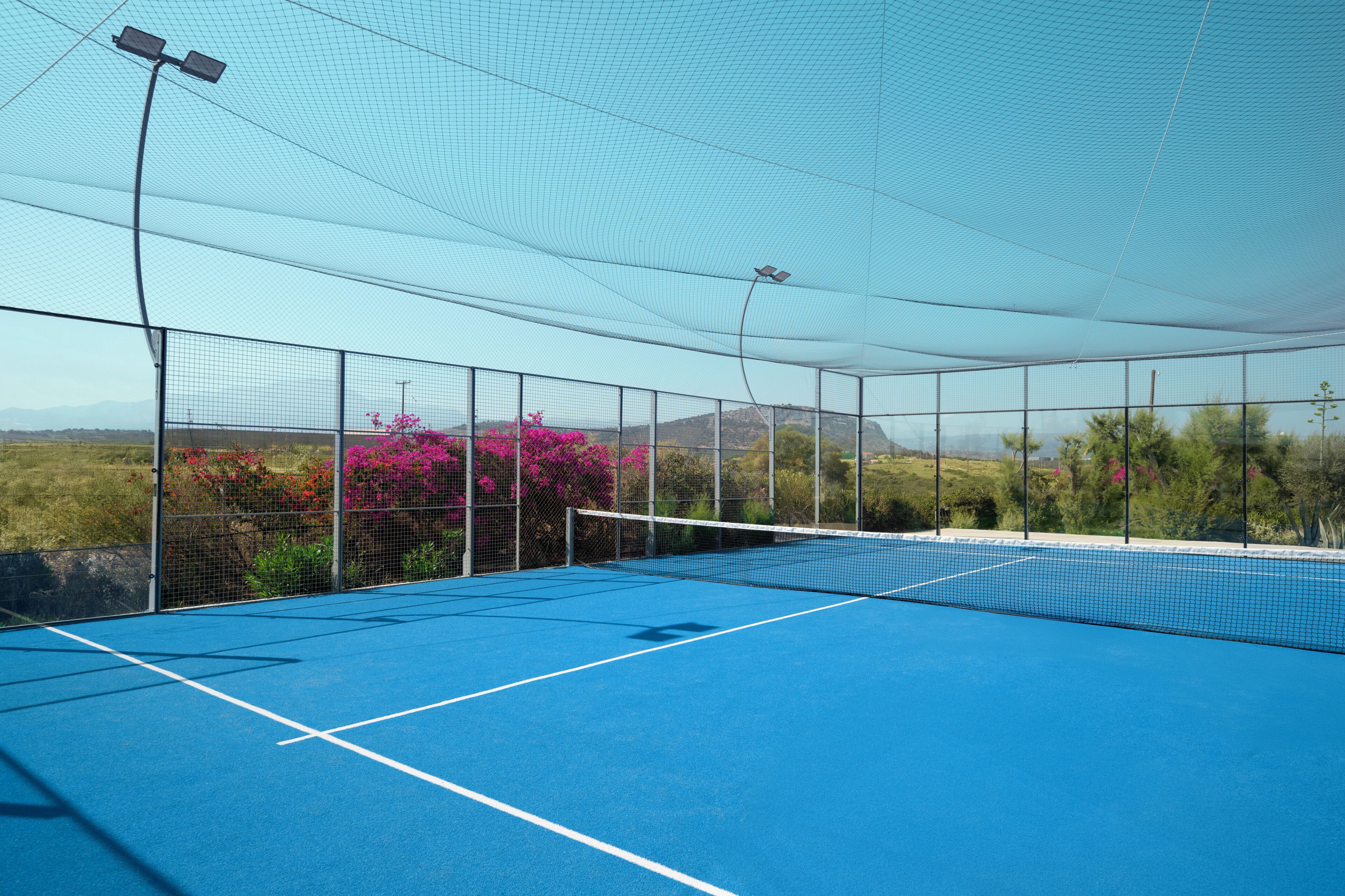 Een buitenterrein voor tennis met een net, omgeven door een hek en overdekt met een luifel.