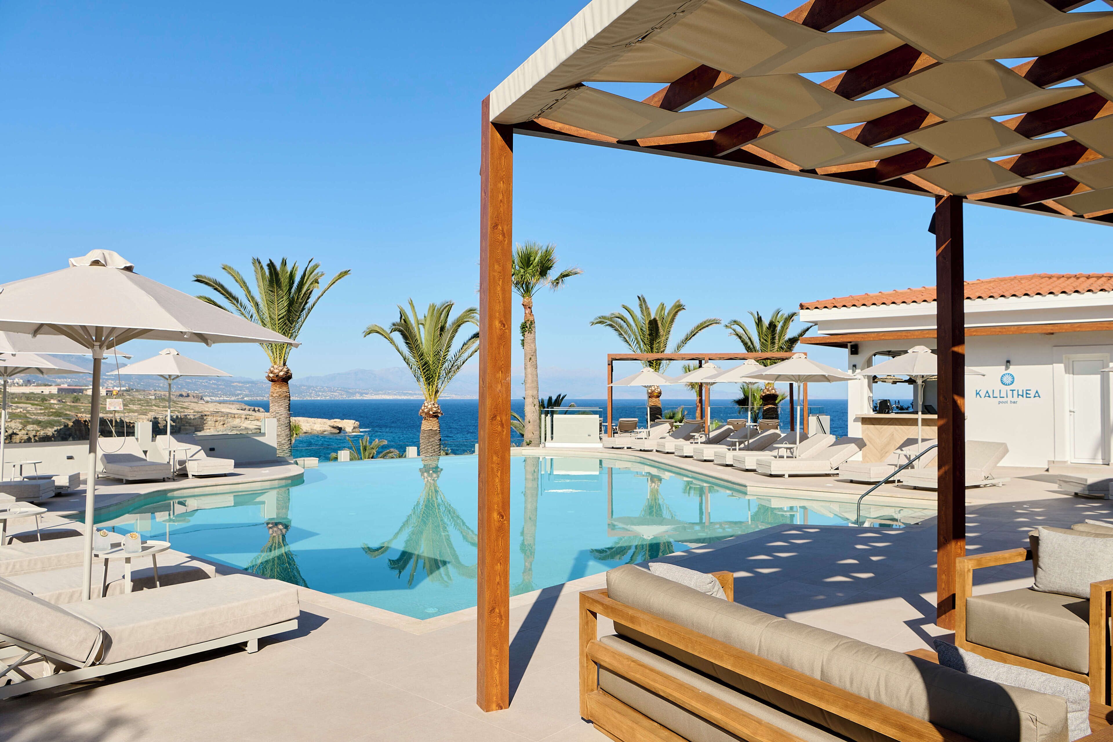 A pool area with lounge chairs and umbrellas, a wooden pergola, and a view of the sea.