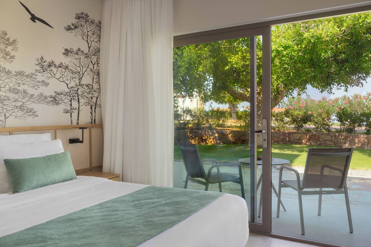 Une chambre d’hôtel avec un lit, un plaid vert, une fresque représentant des arbres et une porte-fenêtre coulissante donnant sur une terrasse extérieure avec des chaises et une table.
