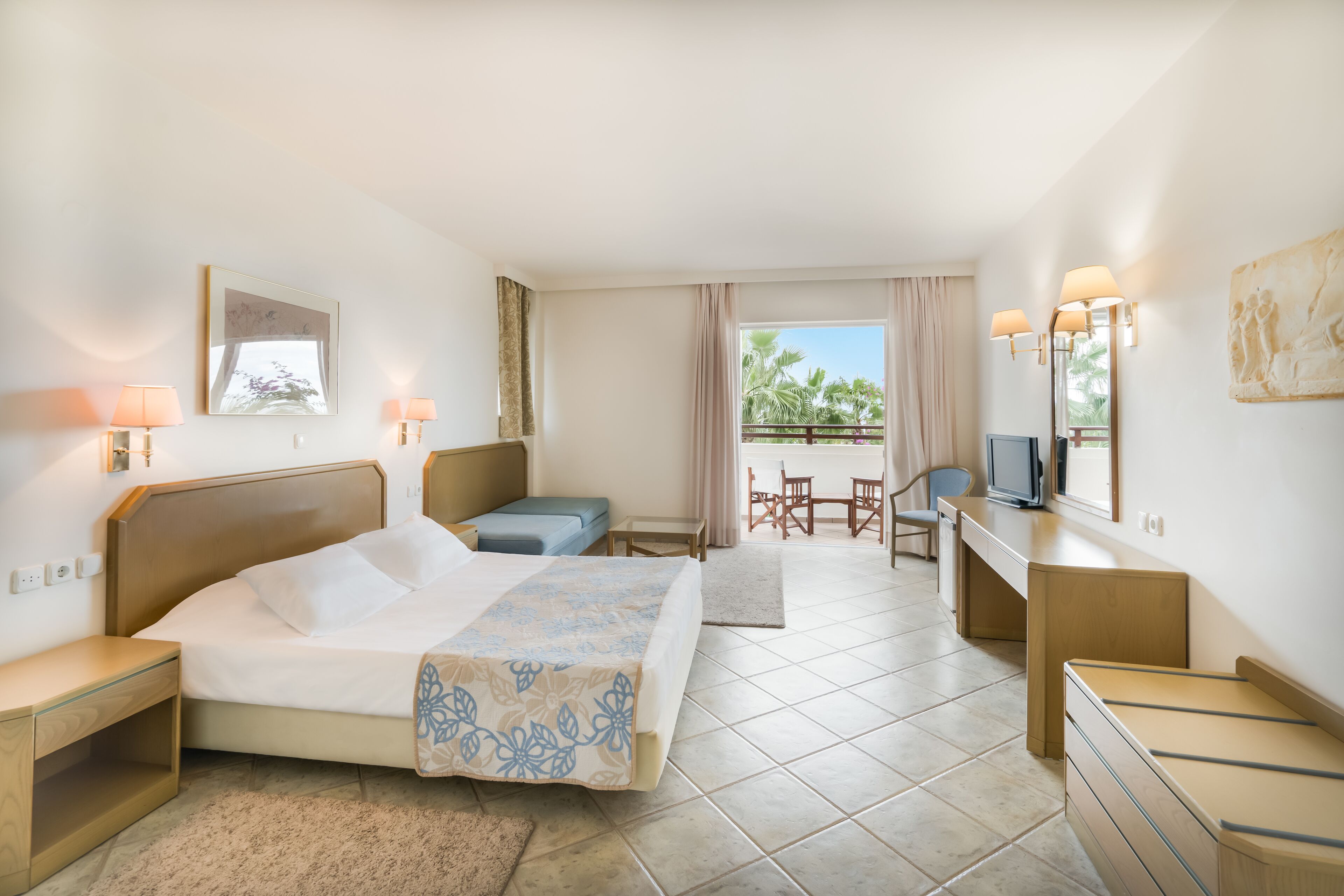 Habitación de hotel con cama, escritorio, silla, televisor y balcón con vista a palmeras.