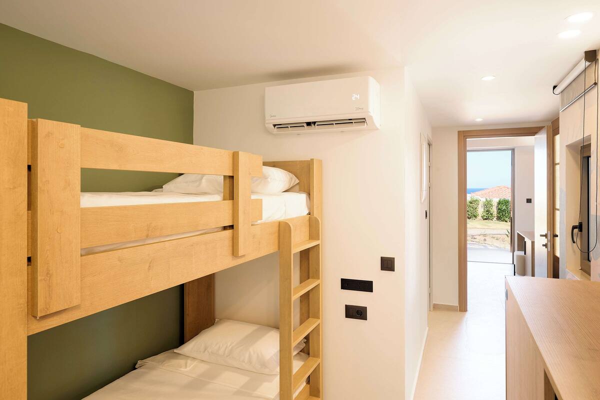 Une chambre avec des lits superposés, une tête de lit en bois, un climatiseur fixé au mur et une vue sur l’extérieur grâce à une porte ouverte.