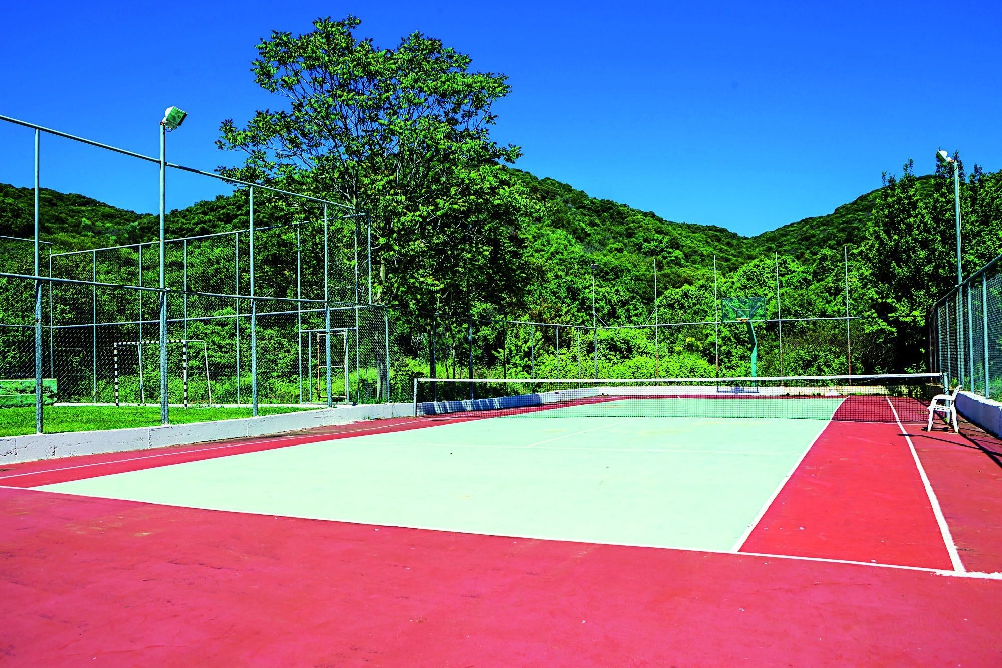Un court de tennis extérieur doté d’une surface de jeu rouge et verte, entouré d’une clôture en treillis métallique et d’arbres.