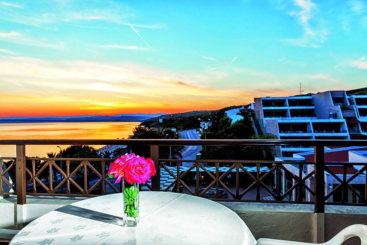 Ein Balkon mit Tisch, einer Blumenvase und Blick auf eine Küstenlandschaft mit Gebäuden.