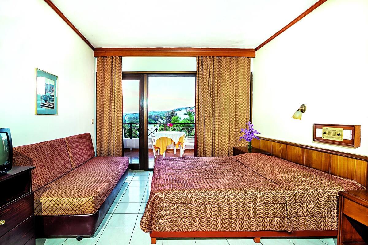 Ein Hotelzimmer mit zwei Betten, einem Fernseher, einem Tisch mit gelber Tischdecke und einem Balkon mit Blick auf die Hügel.