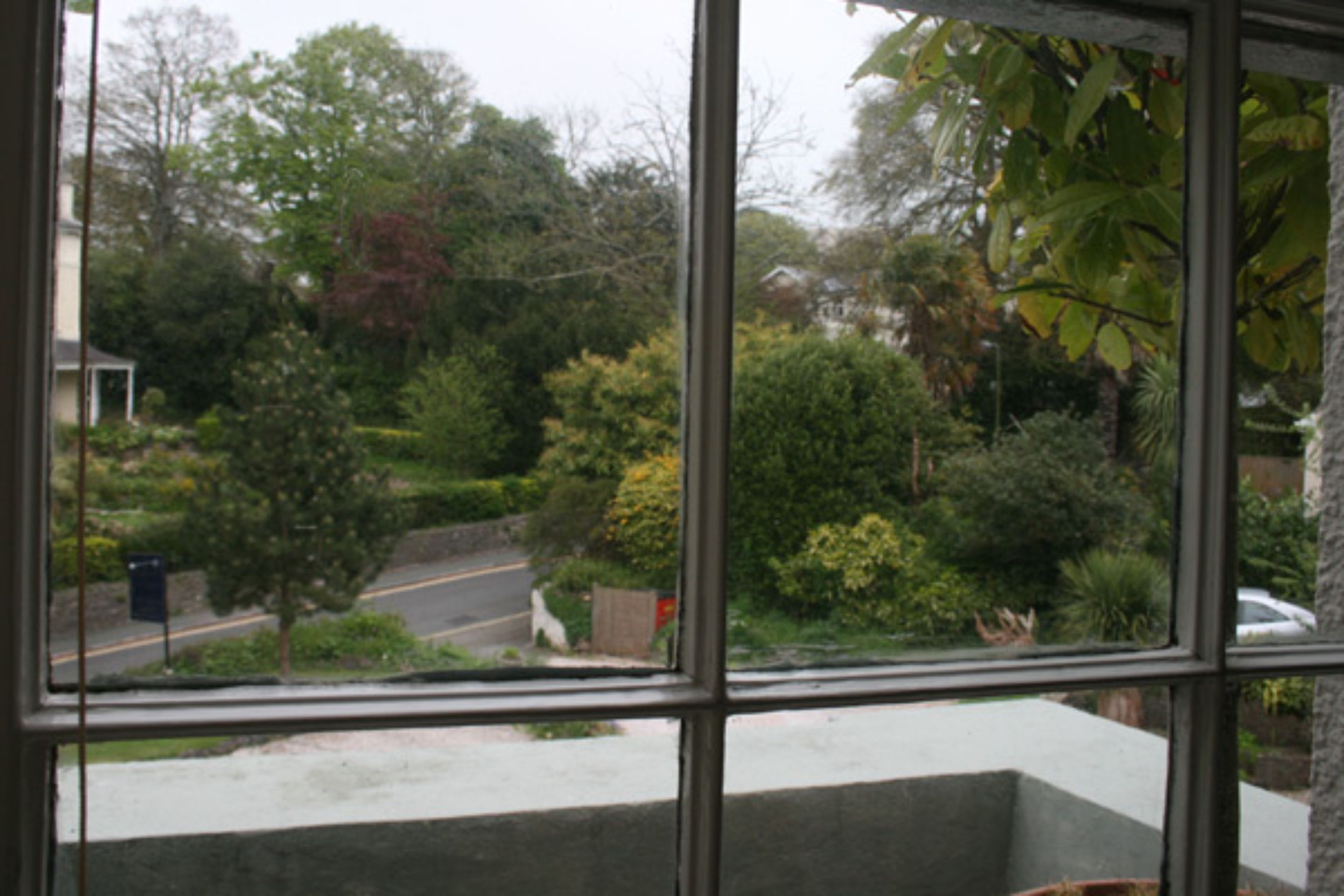 A view through a window with a concrete ledge, trees, and a road.