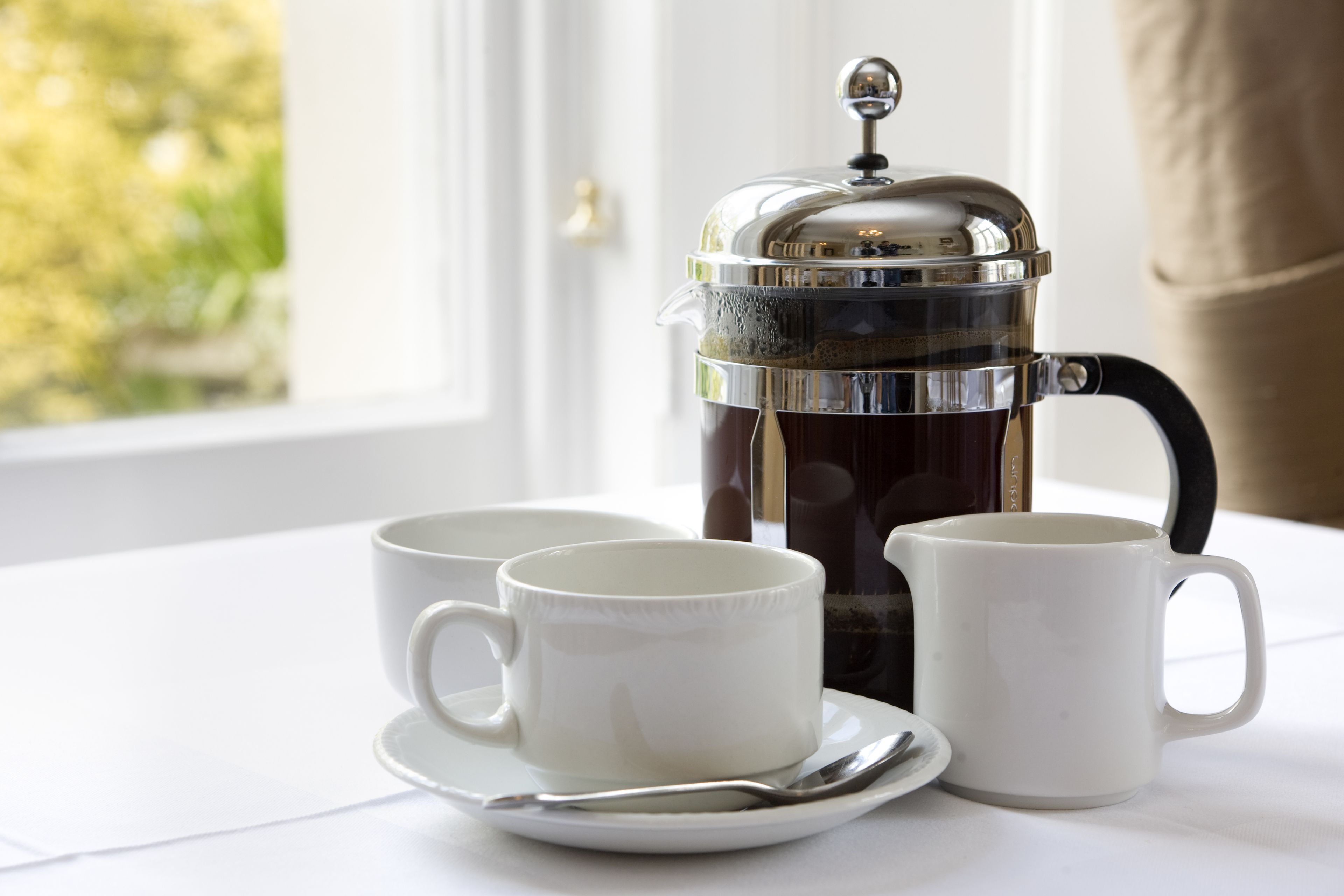 A French press coffee maker on a table with two white cups and a saucer.