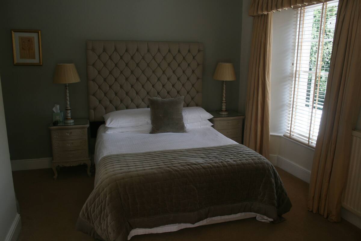 A bedroom with a large bed, two bedside tables, a headboard, and a window with curtains.
