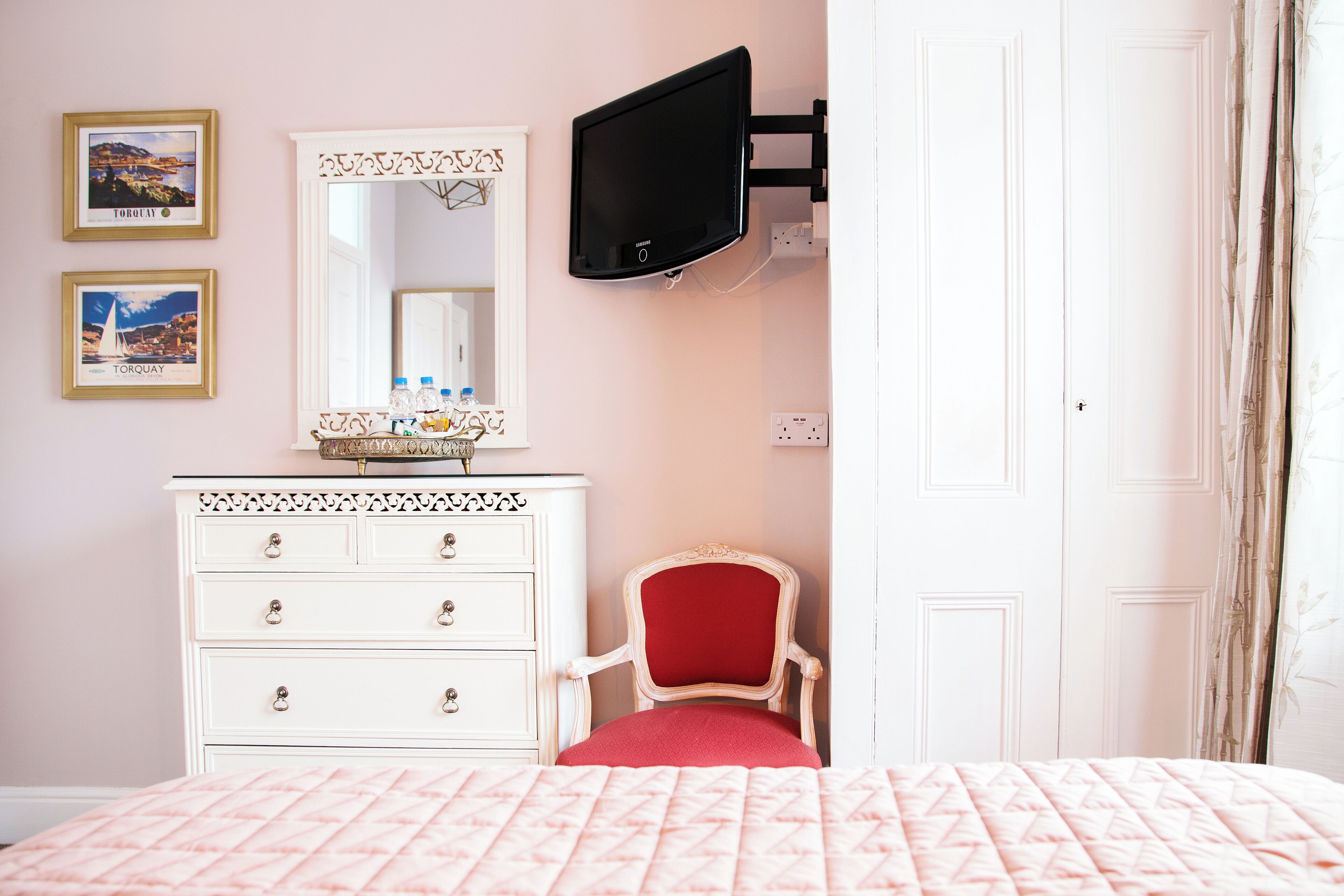 Quarto com cama, cómoda, cadeira, TV fixada na parede e duas imagens emolduradas.
