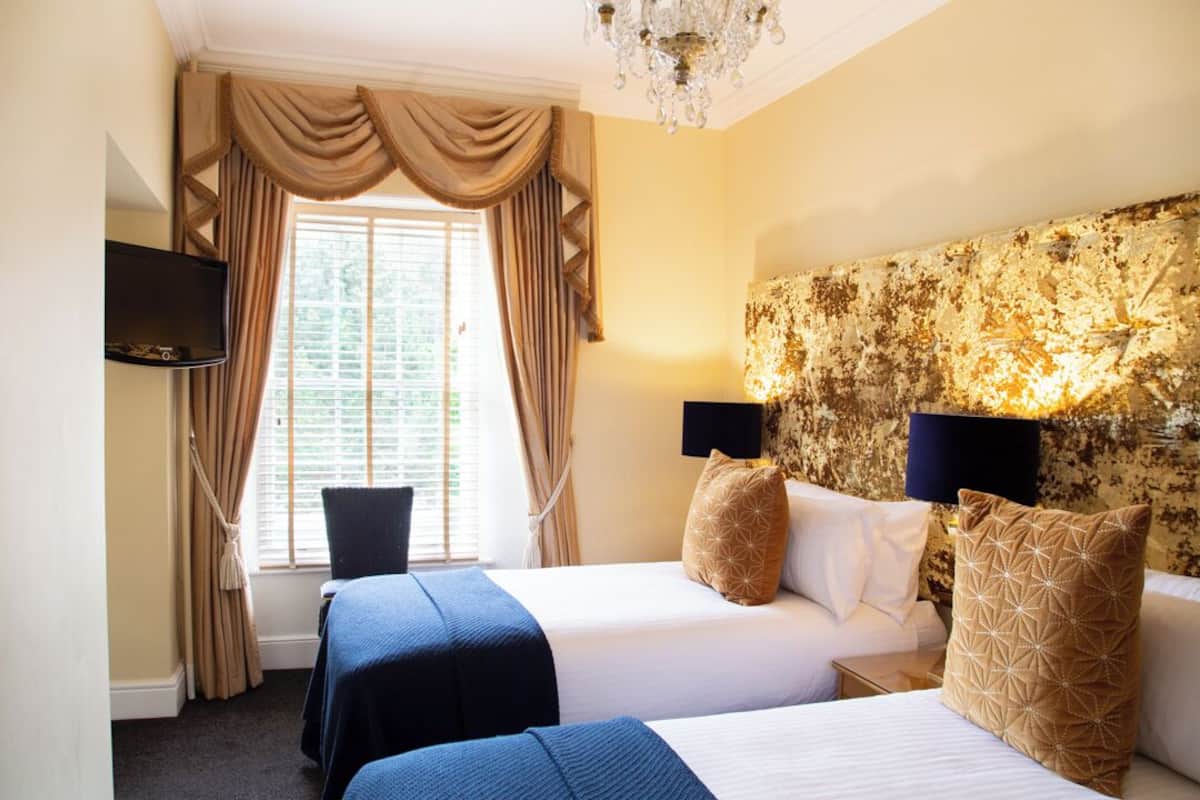 A hotel room with two beds, a TV, a chandelier, and a window with curtains.