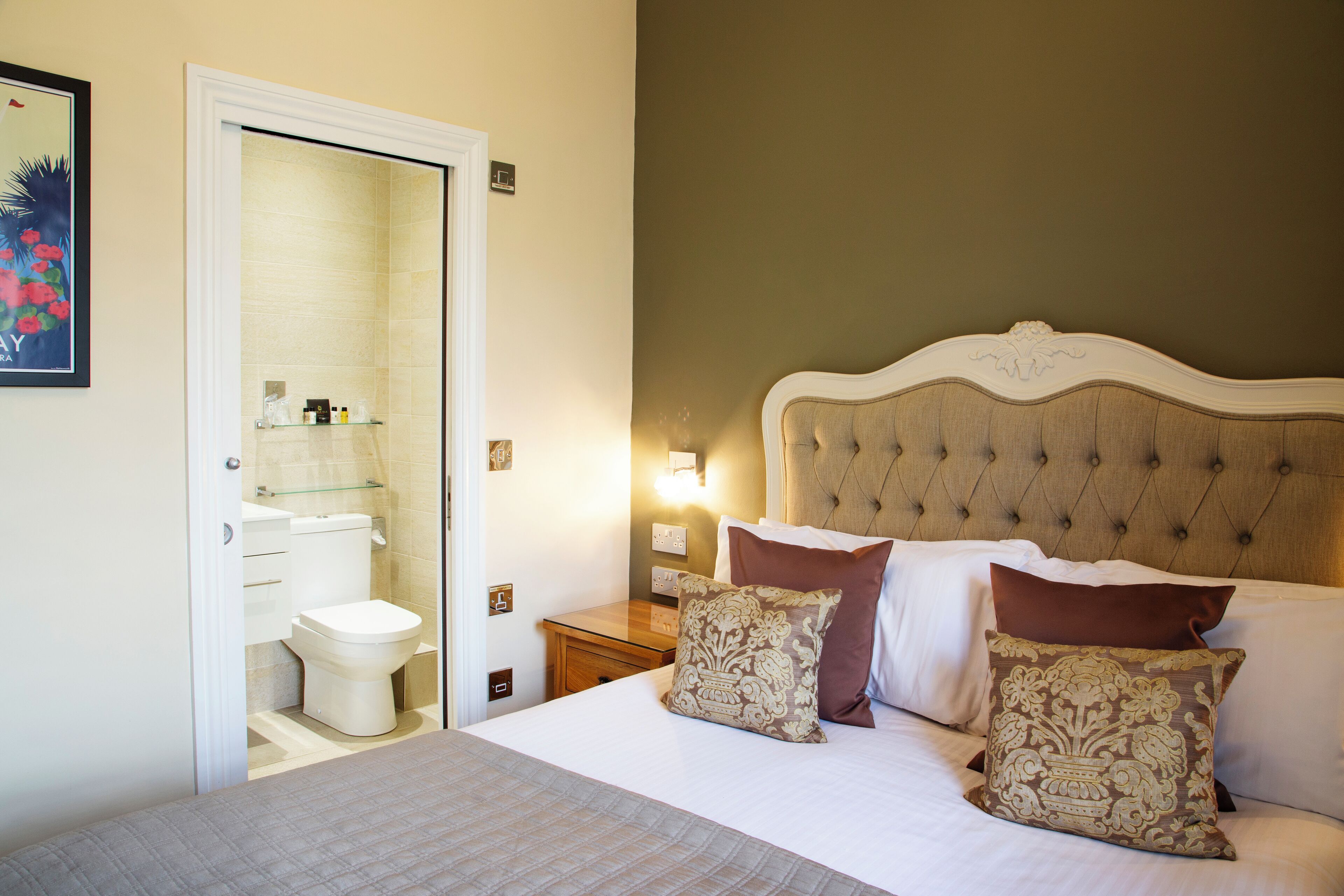 Une chambre à coucher avec un grand lit, une table de chevet, une commode et une salle de bain visible à travers une porte ouverte.