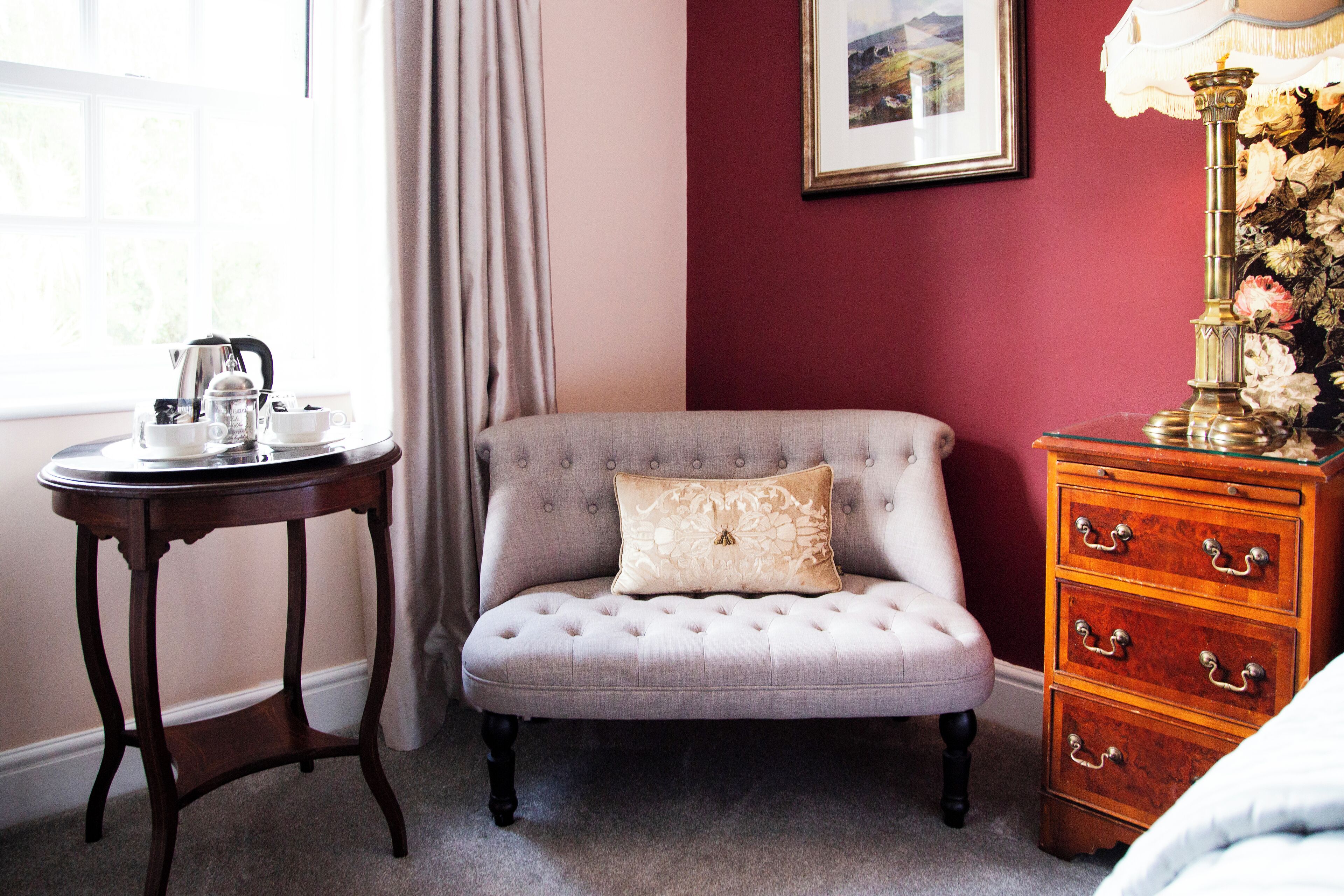 A room with a tufted armchair, a small round table with a teapot, a dresser with drawers, a window with curtains, and a framed picture on the wall.