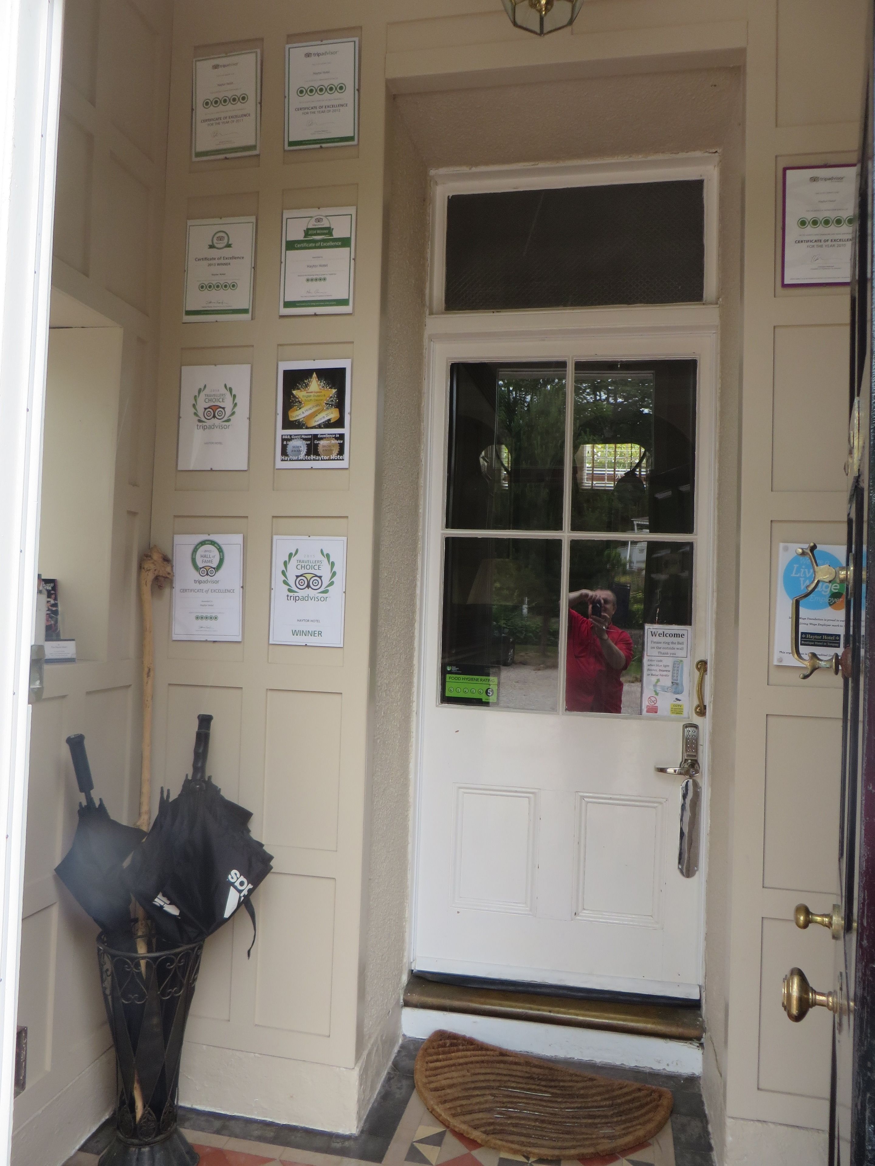 Una puerta con múltiples certificados de excelencia en la pared, un paraguas negro apoyado contra un atril metálico y un felpudo en la entrada.