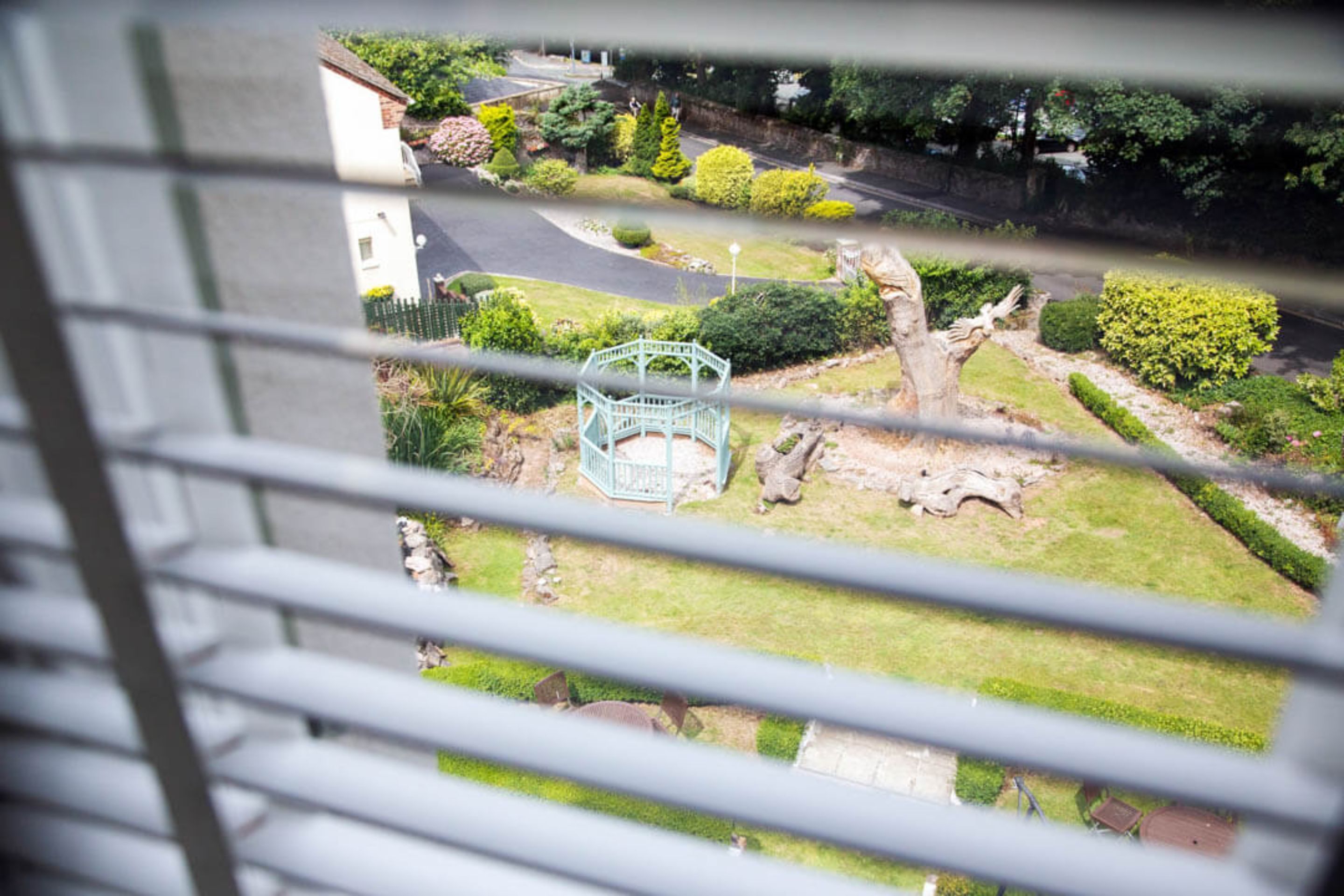 A view through window blinds of a residential area with a small park, a gazebo, and a road.
