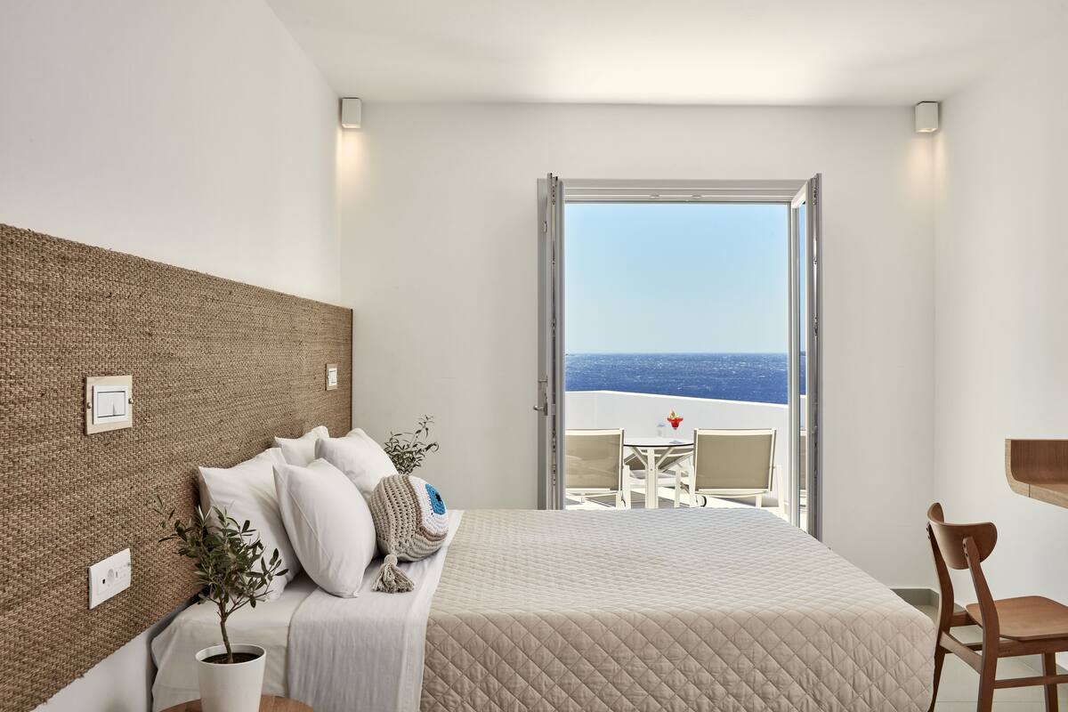 Una habitación de hotel moderna con una cama grande, vista al mar, un balcón con mesa y sillas, y un escritorio de madera.
