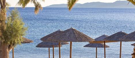 Ein Strand mit Holzliegen und Reetdachschirmen mit Blick auf ein ruhiges Meer und entfernte Berge.