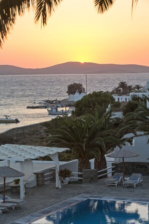 A pool area with lounge chairs, a pergola, and a view of the sea at sunset. - Manoulas Beach Mykonos Resort (Mykonos)