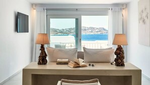 A bedroom with a large window offering a view of the sea and hills. There are two bedside lamps, a bed with white bedding, and a small table with books and glasses.