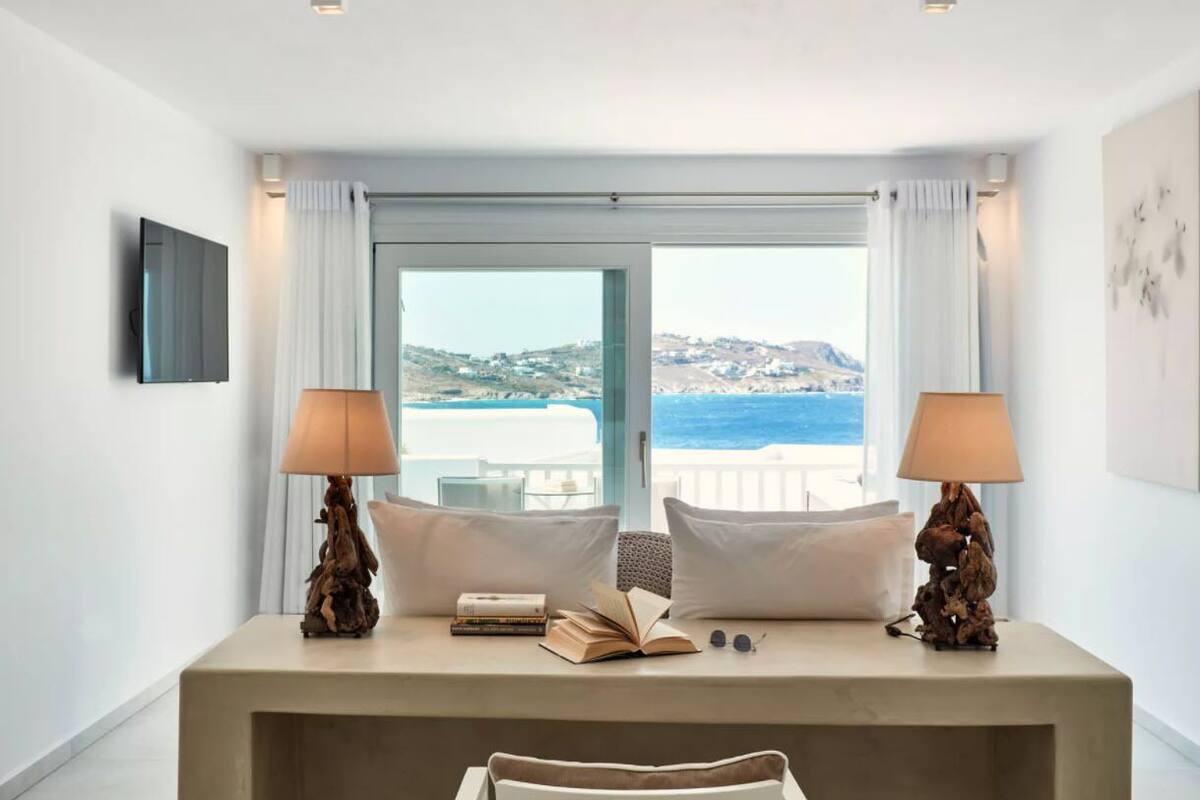 Un dormitorio con un gran ventanal que ofrece vistas al mar y a las colinas. Hay dos lámparas de noche, una cama con ropa de cama blanca y una mesita con libros y vasos.