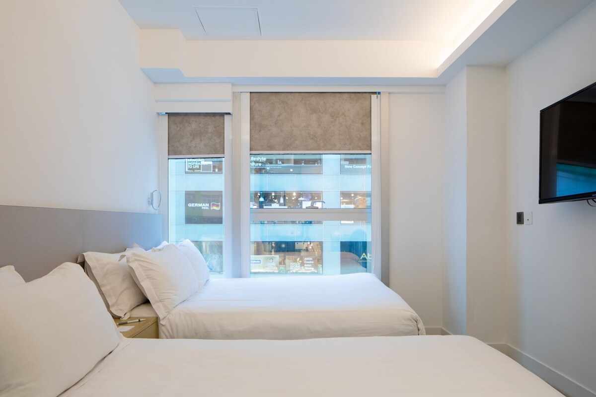 A hotel room with a bed, a TV mounted on the wall, and a view of a shopping mall through the window.