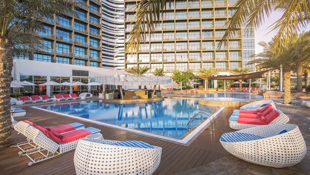 A hotel pool area with lounge chairs, a bar, and a large building in the background.