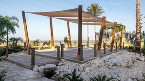 A beachside pavilion with a wooden frame and a canopy, surrounded by palm trees and a rocky beach area.