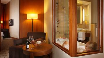 Relax in a spacious room with a seating area, featuring a table and a wine bottle, and enjoy a view of the bathroom with a bathtub and shower.