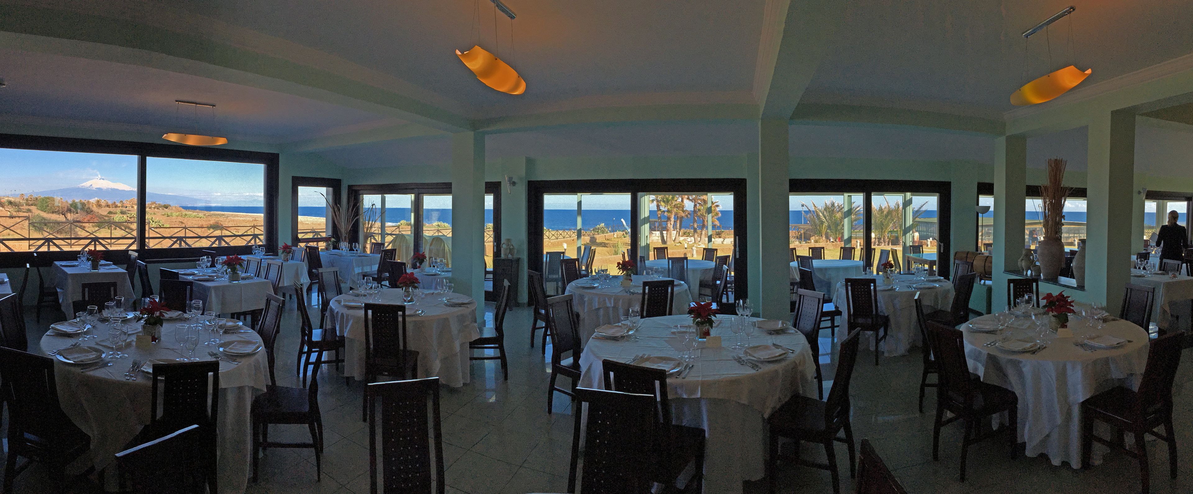A restaurant with multiple round tables set for dining, large windows offering a view of the landscape, and a high ceiling with hanging lights.