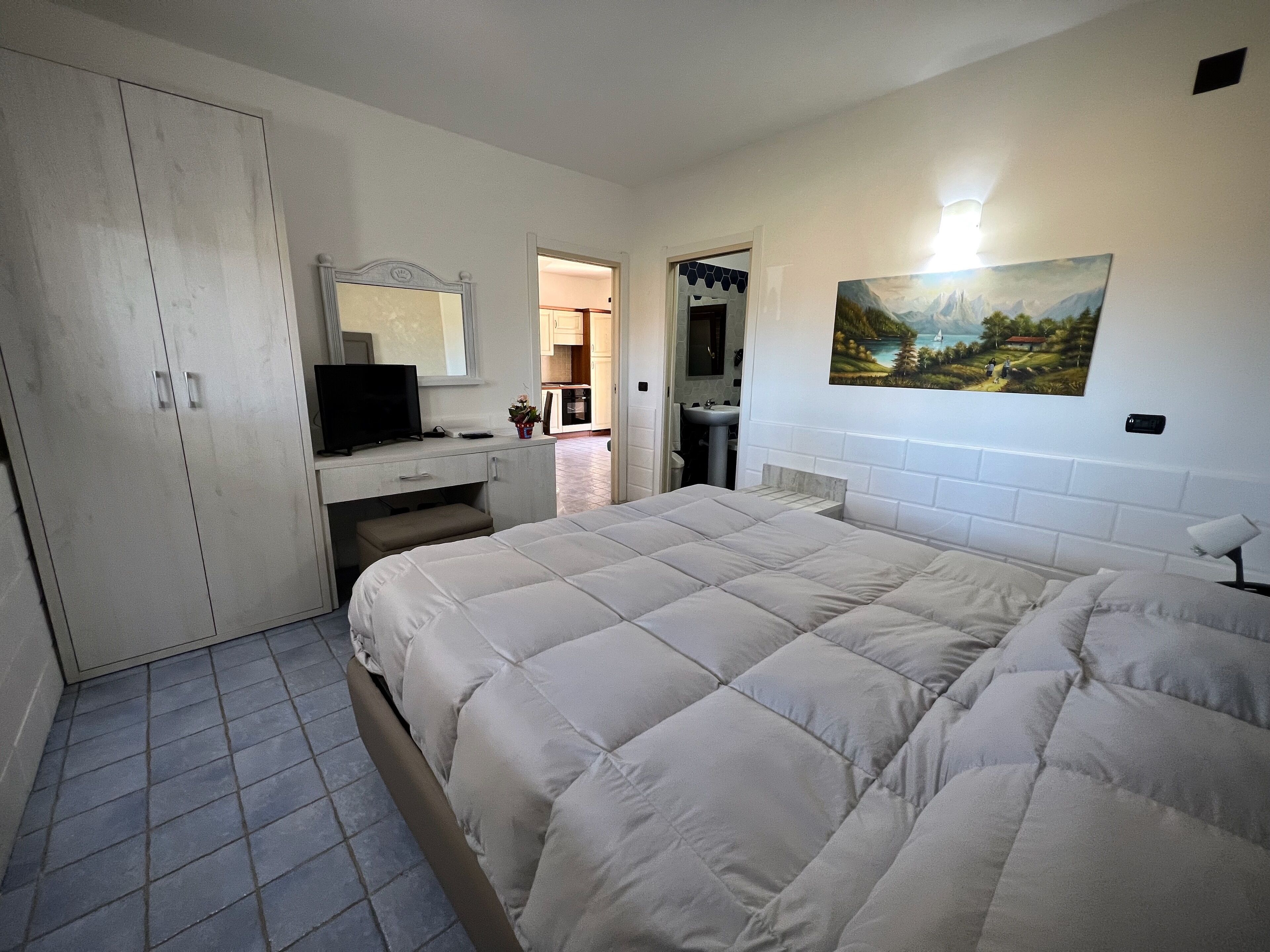A bedroom with a bed, a wardrobe, a desk with a television, a sink, and a painting on the wall.
