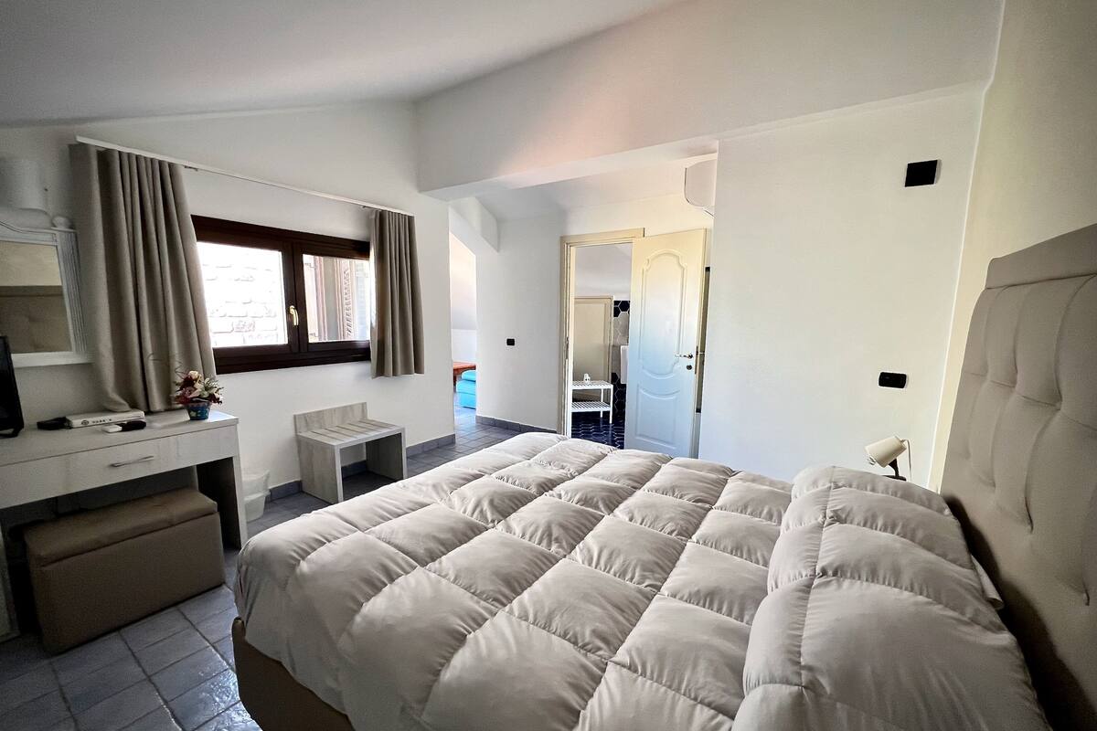 A hotel room with a large bed, a desk with a chair, a small table, and a mirror. There is a window with curtains, a door, and a light fixture on the wall.