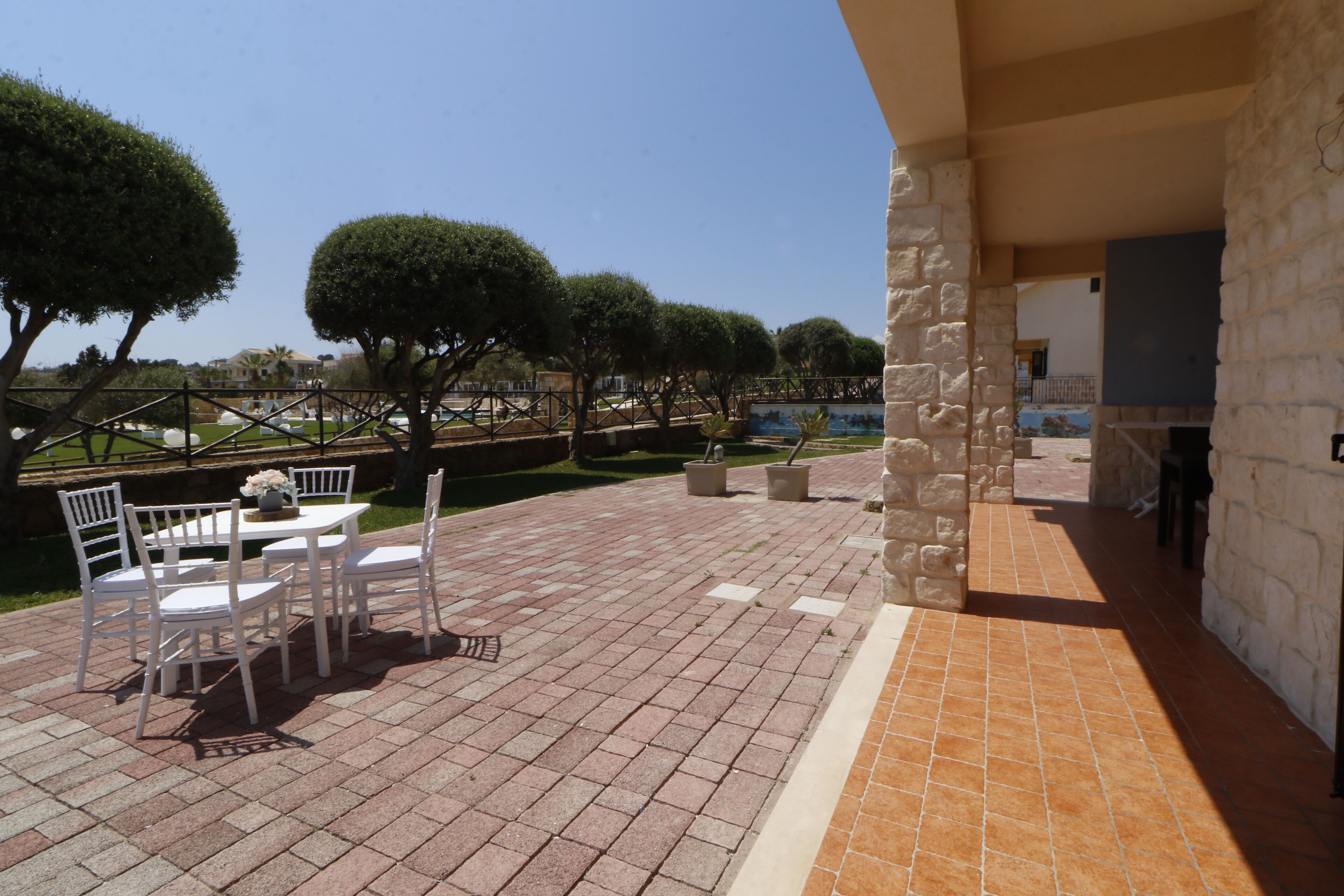 Un patio al aire libre con un muro de piedra, una mesa con sillas y árboles cuidadosamente podados.