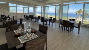 A restaurant with tables set for dining, large windows offering ocean views, and a stone wall. - Hotel Residence Capo Campolato (Augusta)