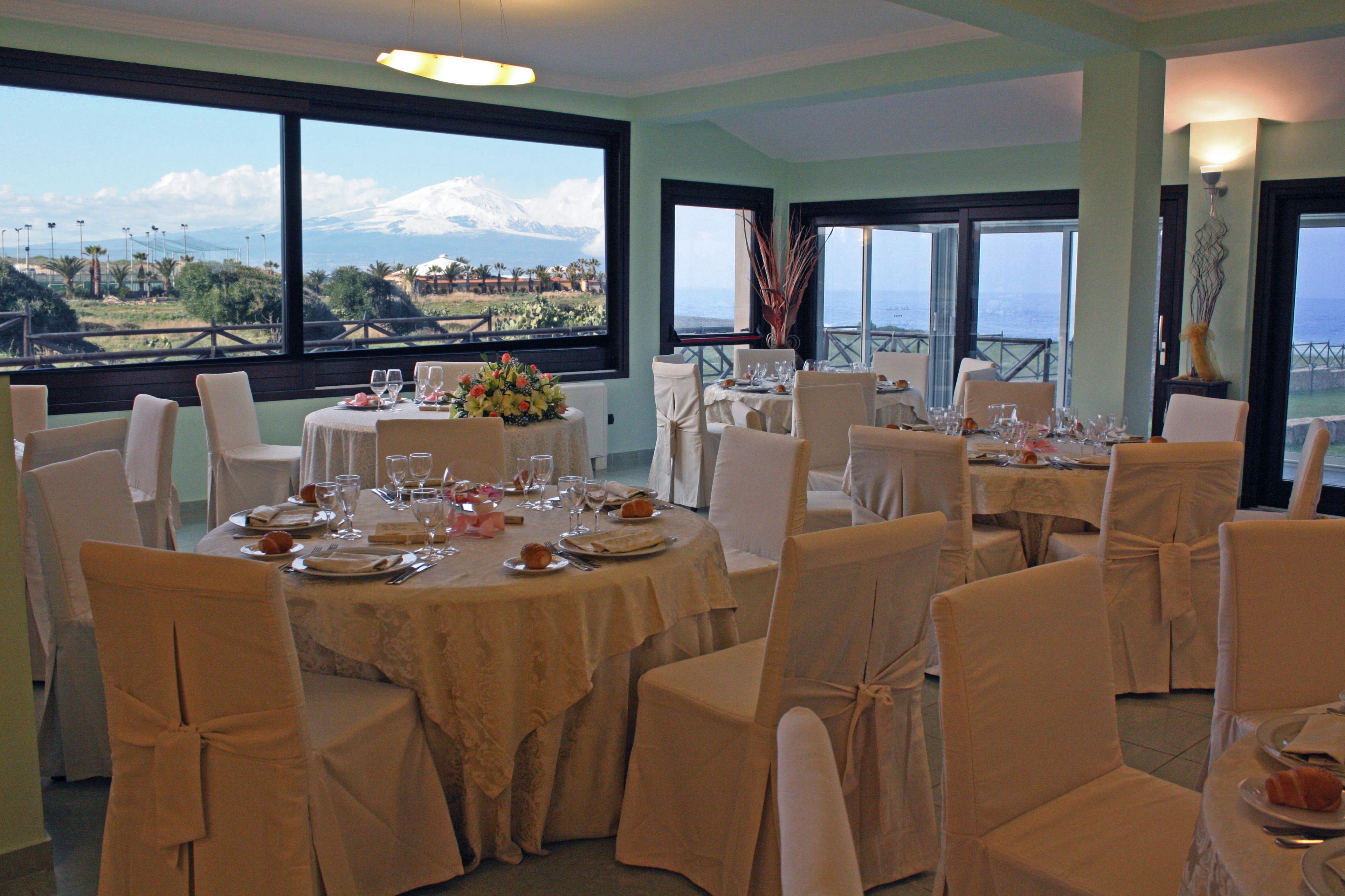 Un comedor con mesas dispuestas para una comida, sillas con fundas blancas y grandes ventanales que ofrecen vistas a un paisaje montañoso.