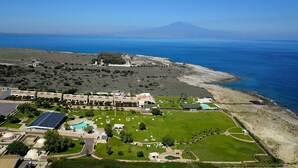 An aerial view of a coastal resort with multiple buildings, a large green lawn, and a view of the sea and a mountain in the distance. - Hotel Residence Capo Campolato (Augusta)