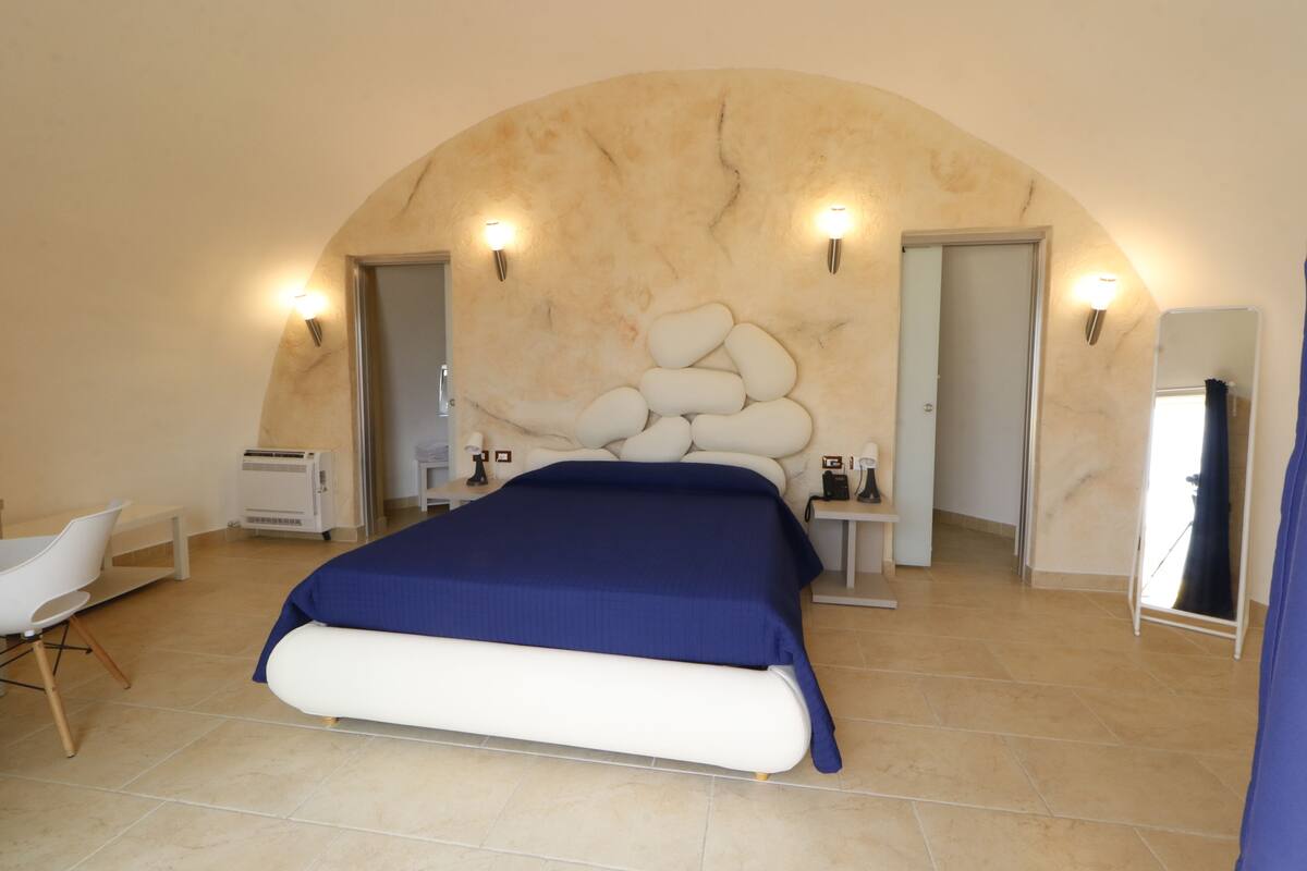 A bedroom with a large bed, a white chair, a small table, and a mirror.
