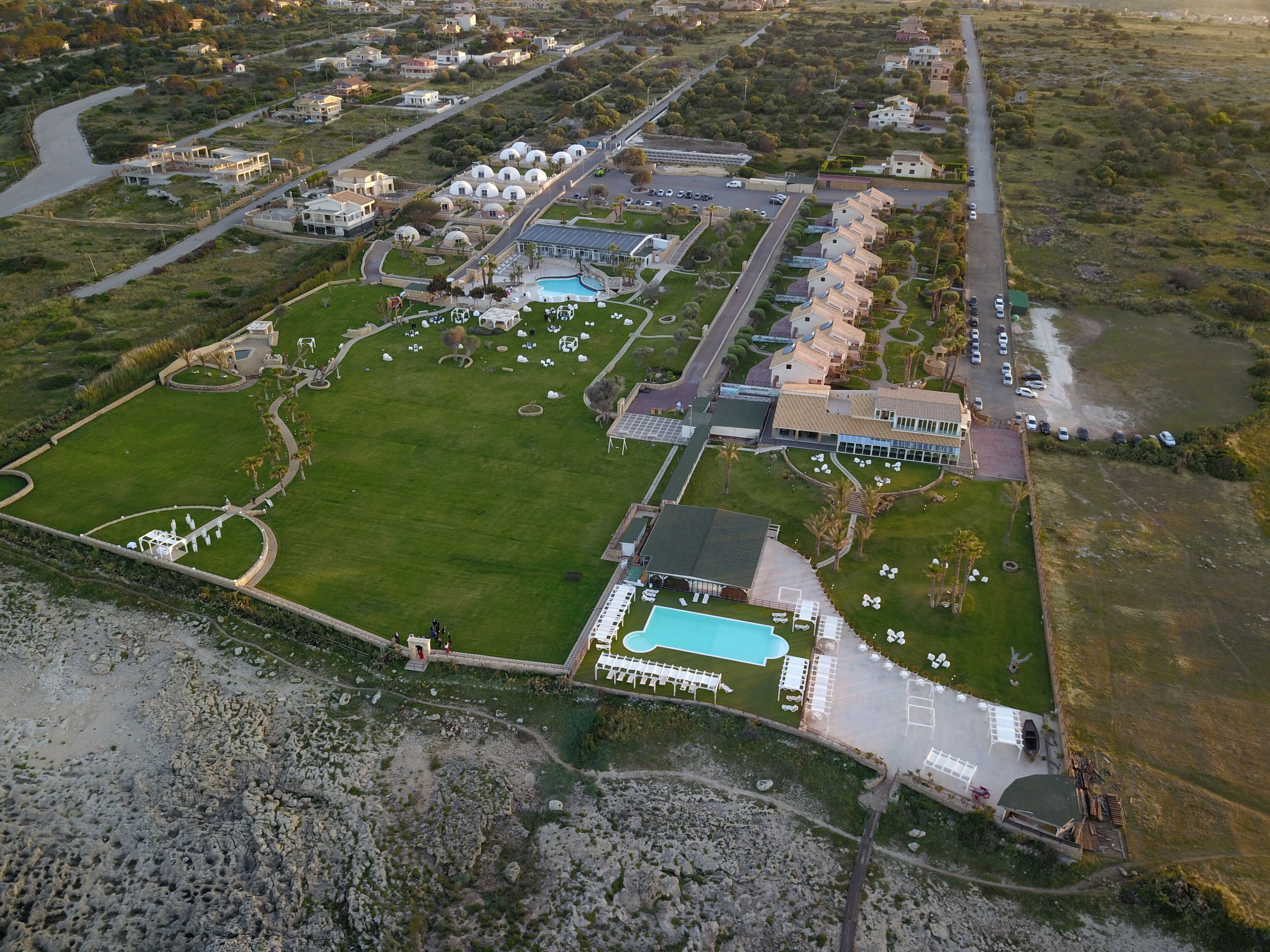 An aerial view of a resort with a large swimming pool, multiple villas, and a central building with a dome structure.