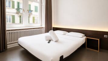 Habitación de hotel con una cama grande, una ventana con cortinas, una mesita de noche y una lámpara de pared.