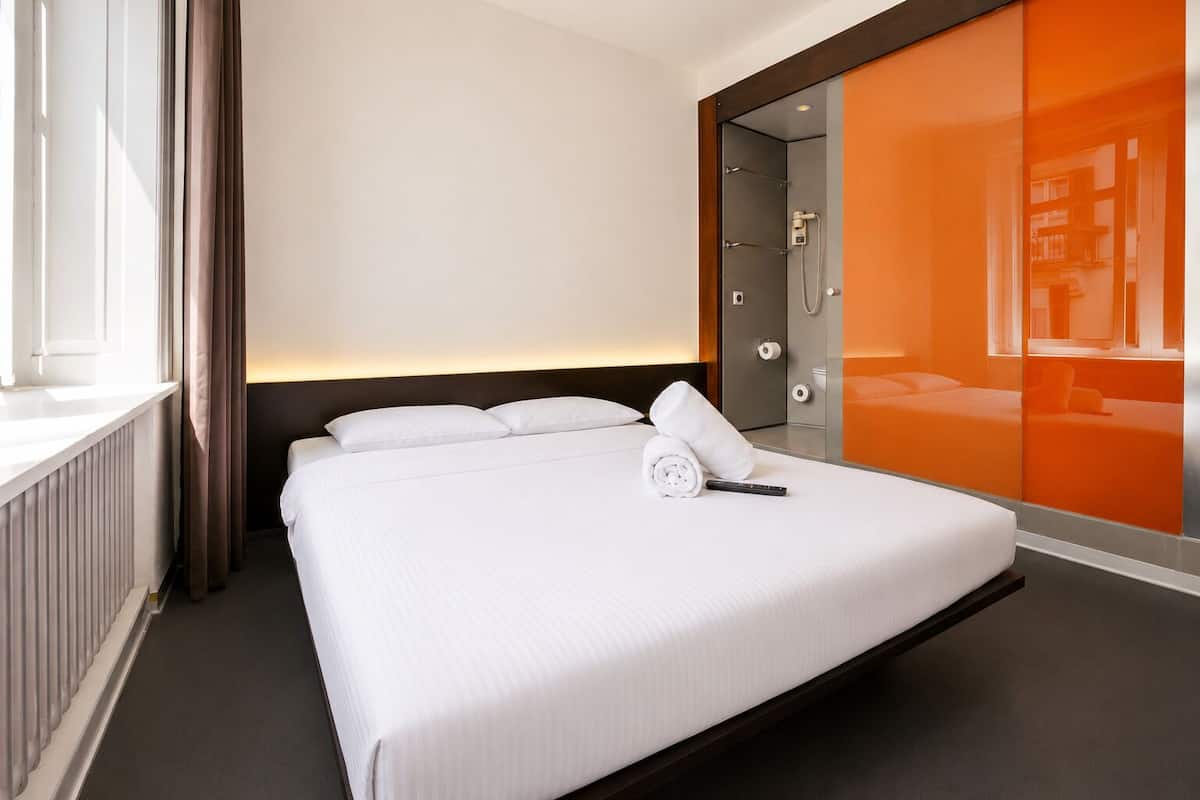 Habitación de hotel con una cama grande, una mesita de noche, una ventana con cortinas, una zona de ducha y una pared de acento color naranja.
