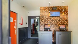 Zona de recepción de un hotel con pared de ladrillo, dos quioscos de auto-registro y un letrero naranja de EasyHotel.